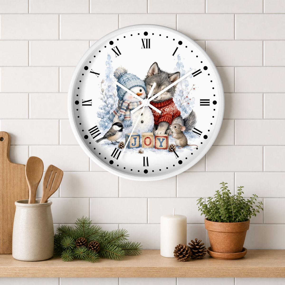 A round white wall clock with a festive winter animal illustration and the word "JOY" in block letters, mounted on a white subway tile wall above a wooden shelf with kitchen decor and a potted plant.