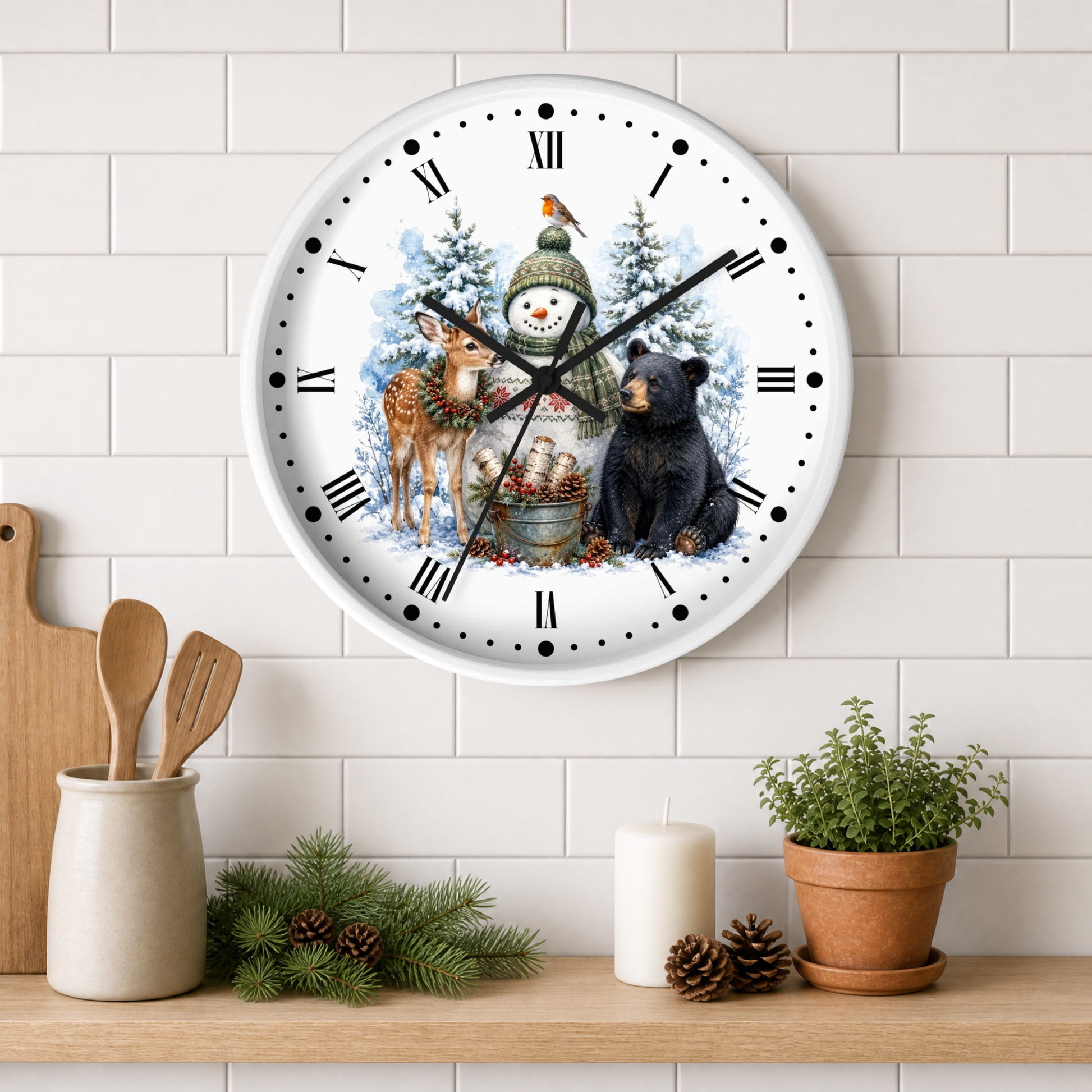 A round wall clock with a white face and black Roman numerals, decorated with a winter holiday illustration featuring a snowman, deer, bear, and evergreen trees. The clock is mounted on a white subway tile wall above a wooden shelf styled with kitchen utensils, pine branches, a candle, and a potted plant.