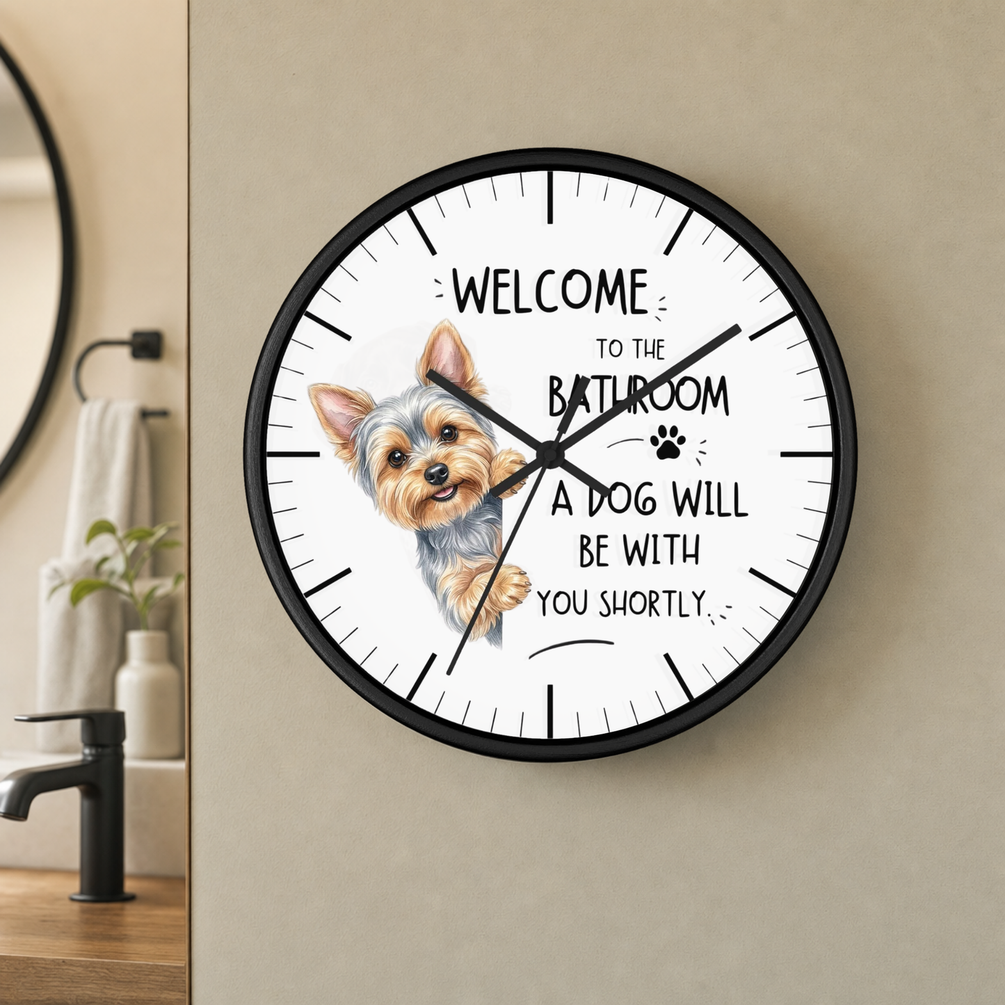 A round wall clock with a black frame and a white face, mounted on a beige wall in a bathroom setting. The clock features a small Yorkshire Terrier illustration and the text 'WELCOME TO THE BATHROOM A DOG WILL BE WITH YOU SHORTLY'.