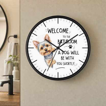 A round wall clock with a black frame and a white face, mounted on a beige wall in a bathroom setting. The clock features a small Yorkshire Terrier illustration and the text 'WELCOME TO THE BATHROOM A DOG WILL BE WITH YOU SHORTLY'.