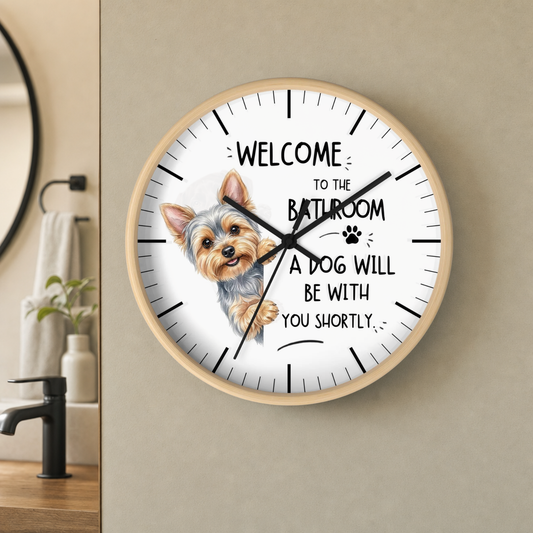 A round wall clock with a light wood frame hangs on a beige wall in a bathroom setting. The clock face is white with black text reading 'WELCOME TO THE BATHROOM A DOG WILL BE WITH YOU SHORTLY' and a printed illustration of a small Yorkie dog.