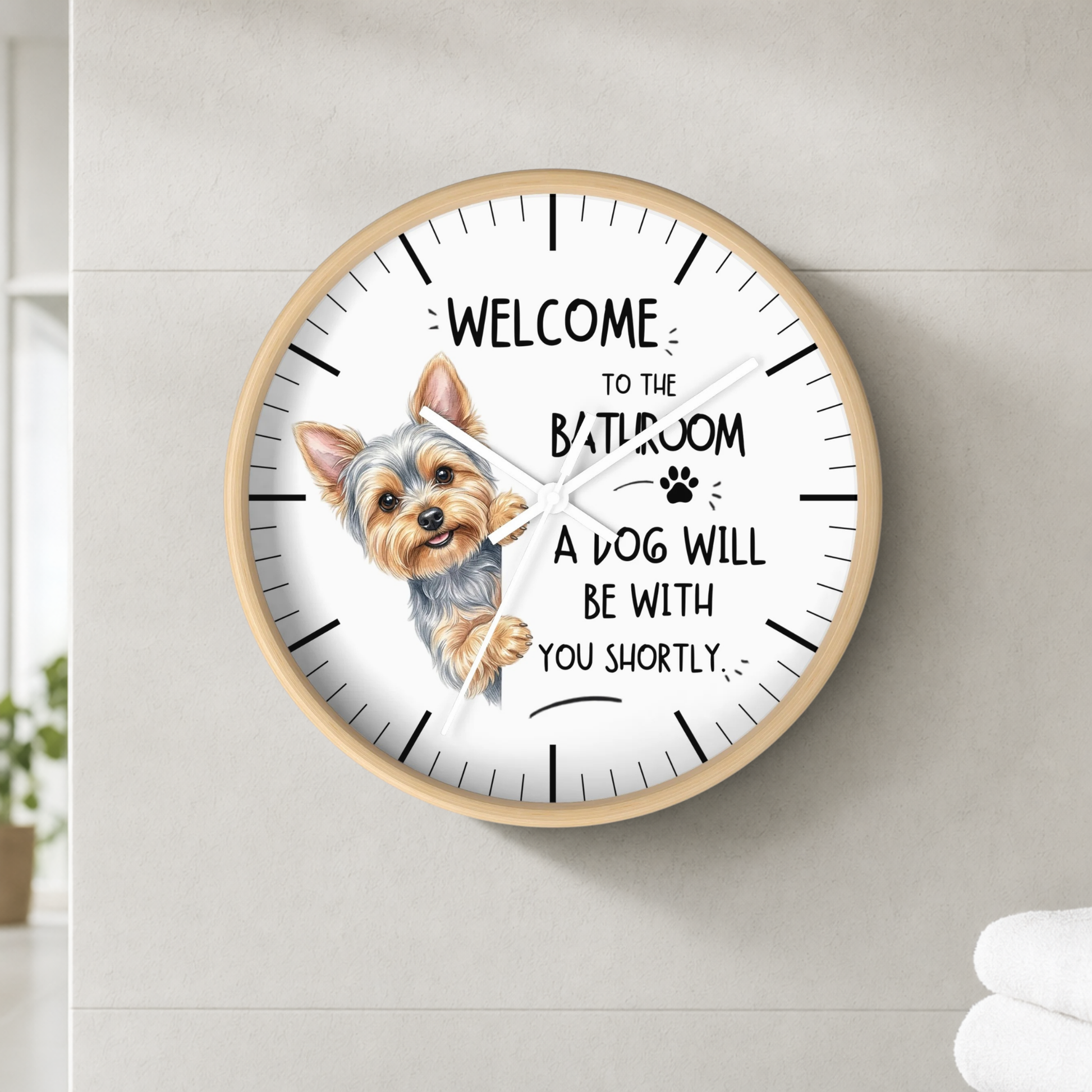A round wall clock with a light wood frame and white face, featuring a small dog illustration and the text 'WELCOME TO THE BATHROOM A DOG WILL BE WITH YOU SHORTLY.' The clock is mounted on a light gray tiled wall.